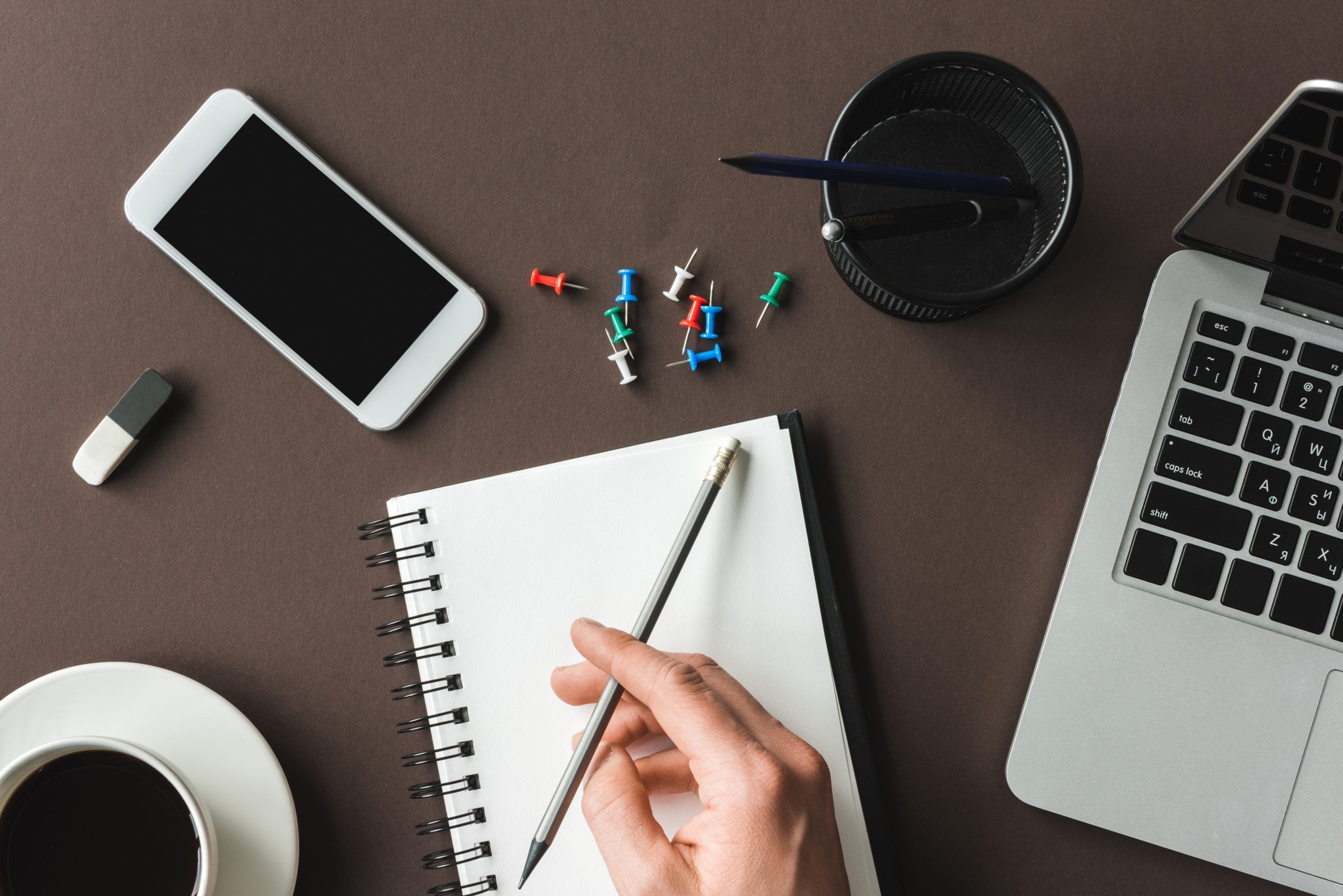 Person holding pencil poised over a notebook on a desk with phone, laptop, coffee and thumbtacks representing content generation.