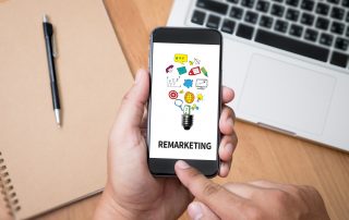 Image showing hands swiping on a smartphone that says "Remarketing" with a laptop and folder in background.