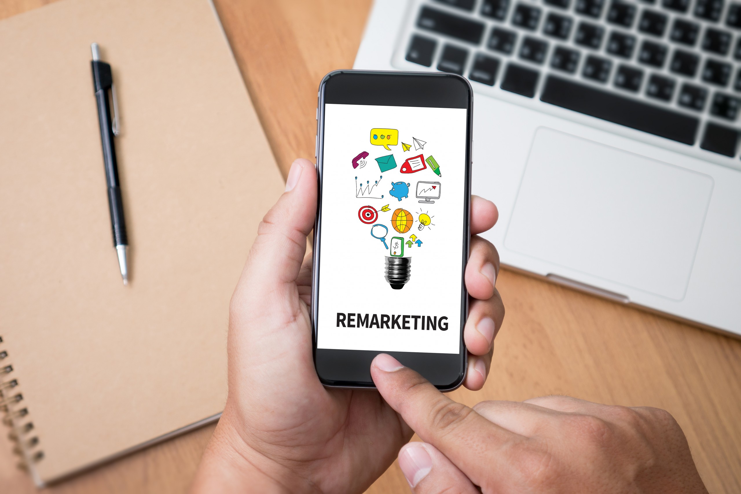 Image showing hands swiping on a smartphone that says "Remarketing" with a laptop and folder in background.