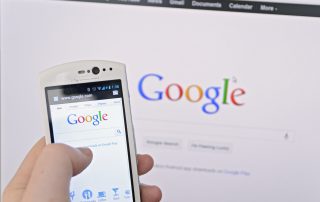 Image of a person on a smartphone conducting a Google search on a phone in front of a computer showing Google on screen.