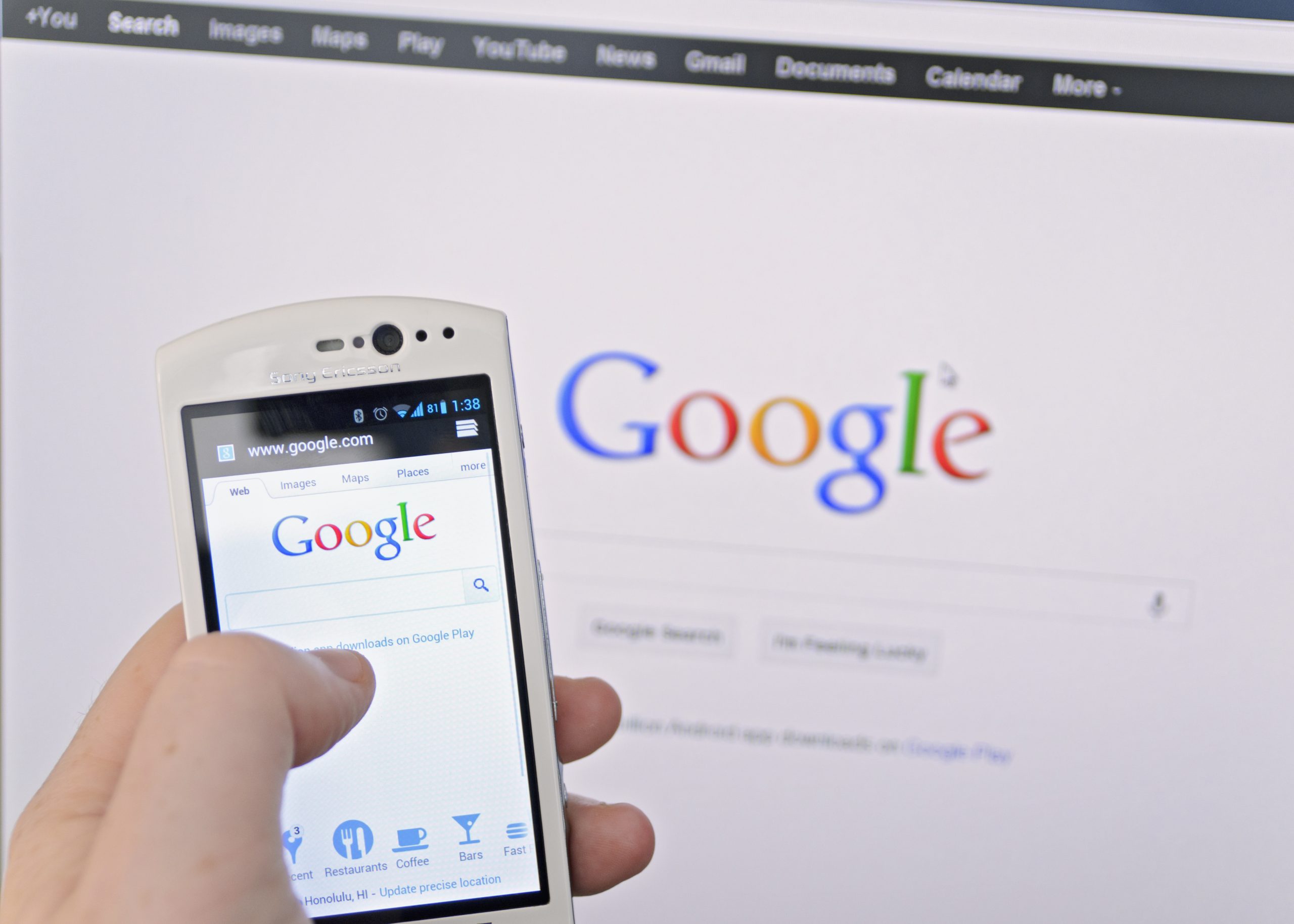 Image of a person on a smartphone conducting a Google search on a phone in front of a computer showing Google on screen.