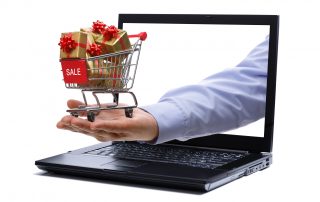 Man's hand holding tiny shopping cart full of holiday gifts through a laptop screen representing holiday digital marketing.