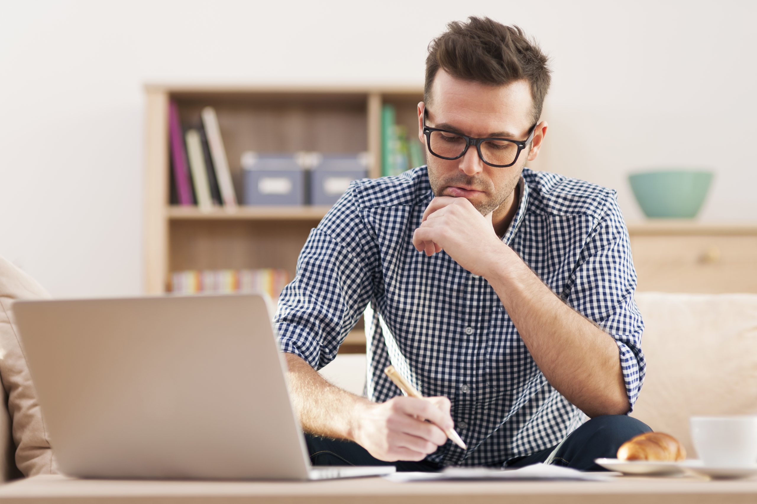Image of a young professional man concentrating in front of a laptop with a pen in his hand, working at home. 