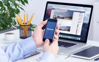 Small business owner at a desk using Facebook on his mobile device and laptop which represents Facebook retargeting.